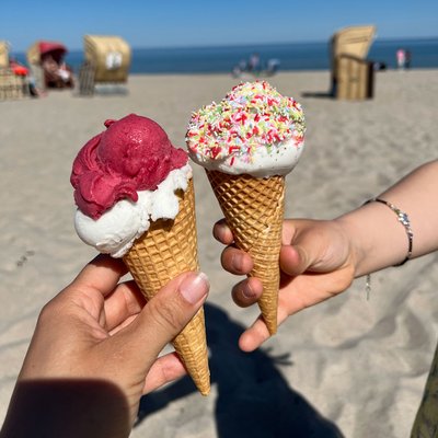 Zwei Personen "stoßen" mit ihren Eiswaffeln an. Im Hintergrund sieht man Strand und blauen Himmel