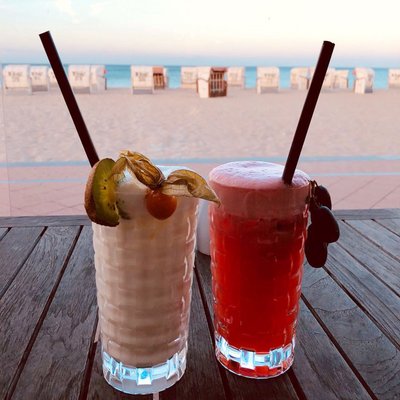 Zwei bunte Cocktails stehen auf einem Holztisch. Im Hintergrund sieht man einen Sandstrand in der Abenddämmerung