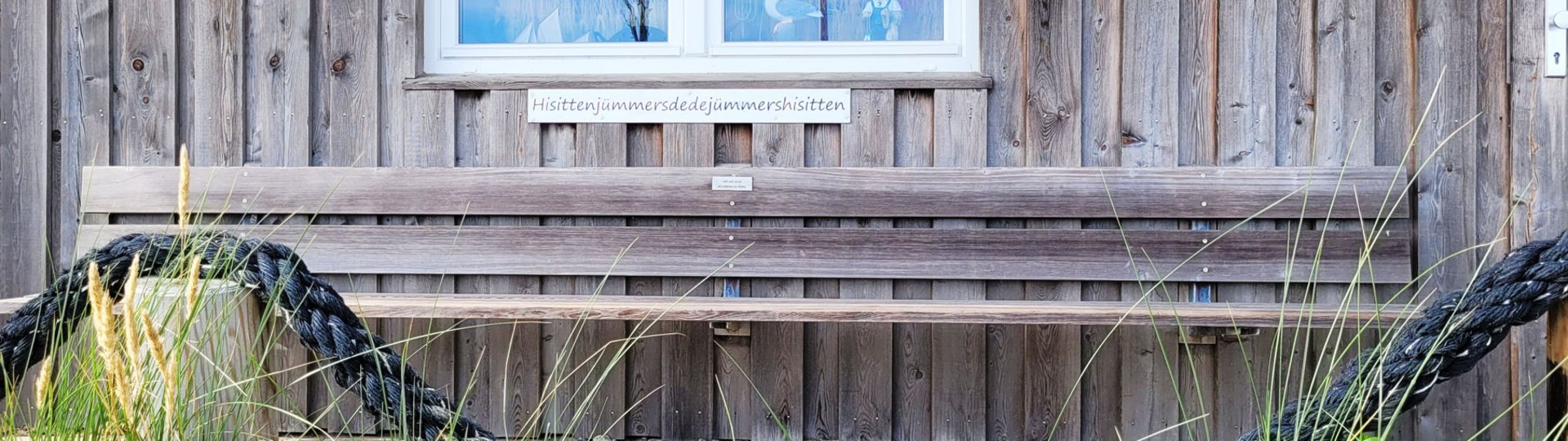 Eine lange Holzbank vor einem Holzhaus. Auf einem Schild steht "Hisittenjümmersdedejümmershisitten". Das ist Plattdeutsch und bedeutet "Hier sitzen immer die, die immer hier sitzen"