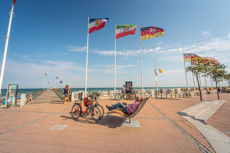 Zwei Radfahrer sitzen auf einem Holzsessel an einer Uferpromenade. Die Räder stehen nebenbei