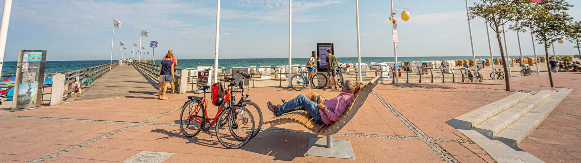 Zwei Radfahrer sitzen auf einem Holzsessel an einer Uferpromenade. Die Räder stehen nebenbei