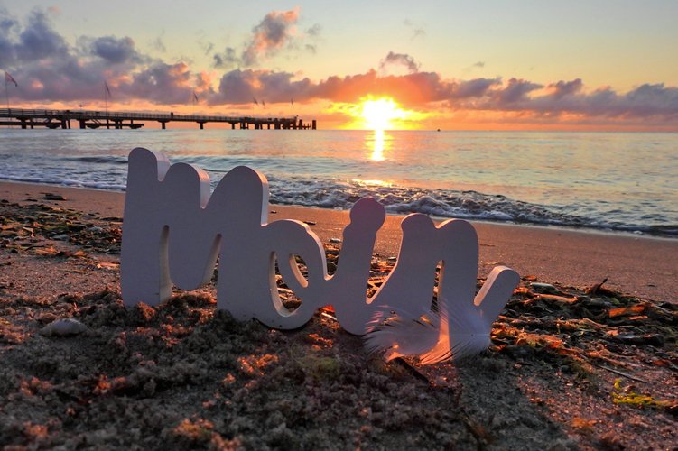 das Wort "Moin" im Sand und die Seebrücke im Hintergrund bei Sonnenuntergang