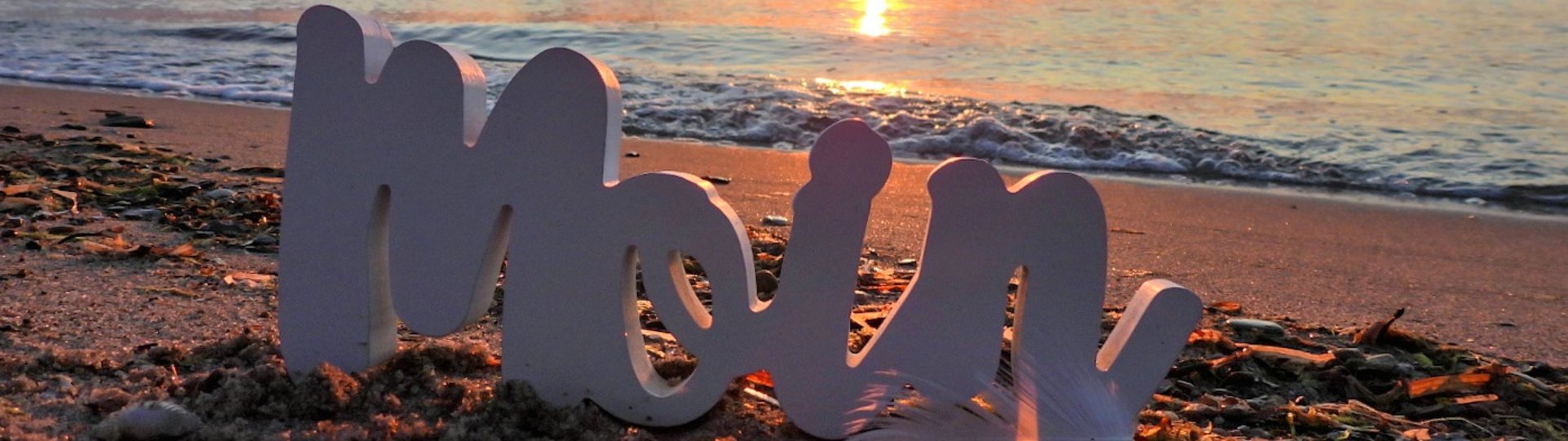 das Wort "Moin" im Sand und die Seebrücke im Hintergrund bei Sonnenuntergang
