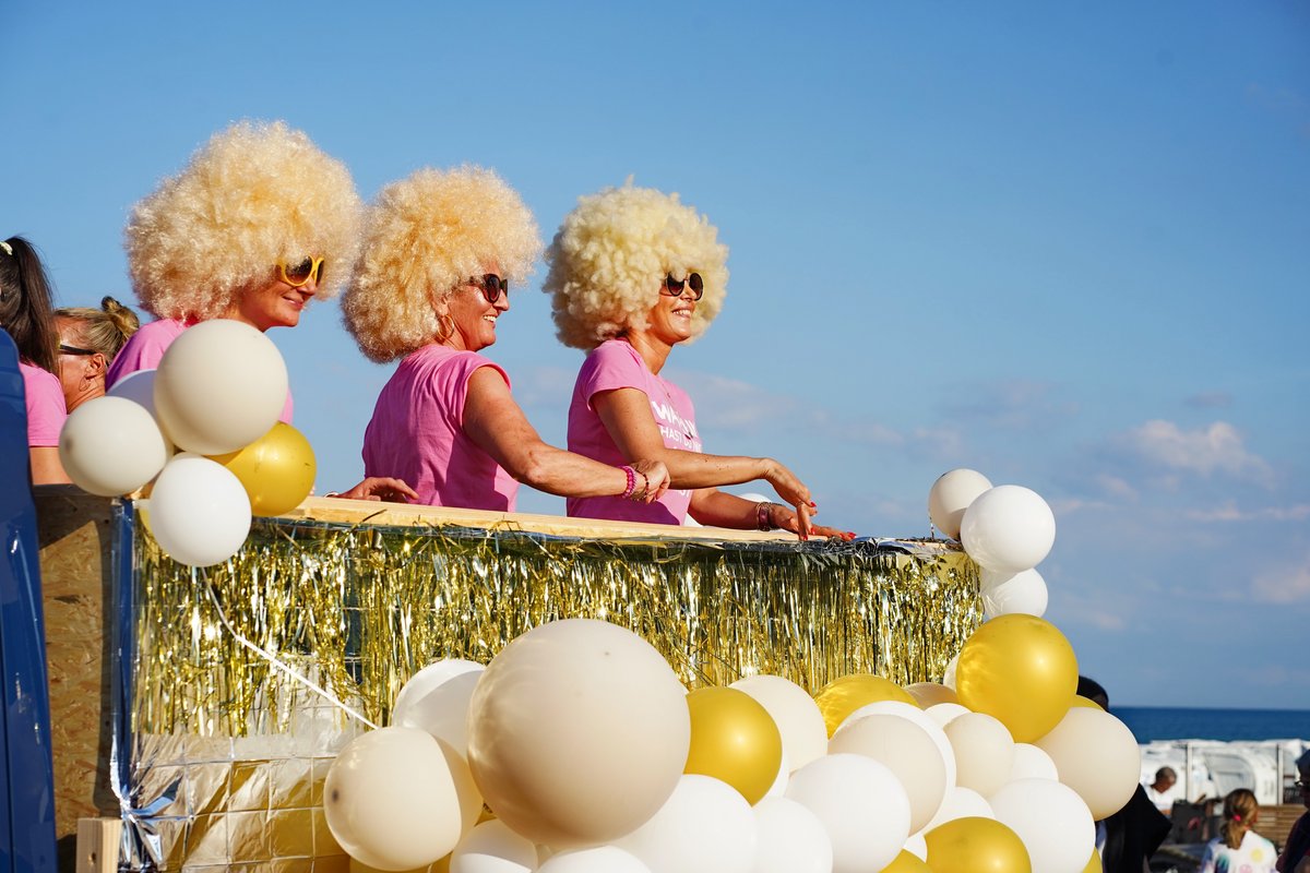 Drei Frauen mit blonden Perücken und rosa T-Shirts stehen fröhlich auf einem Festwagen