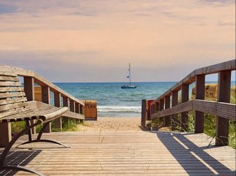 Ein Holzsteg führt an den Strand, am Horizont sieht man an Segelboot