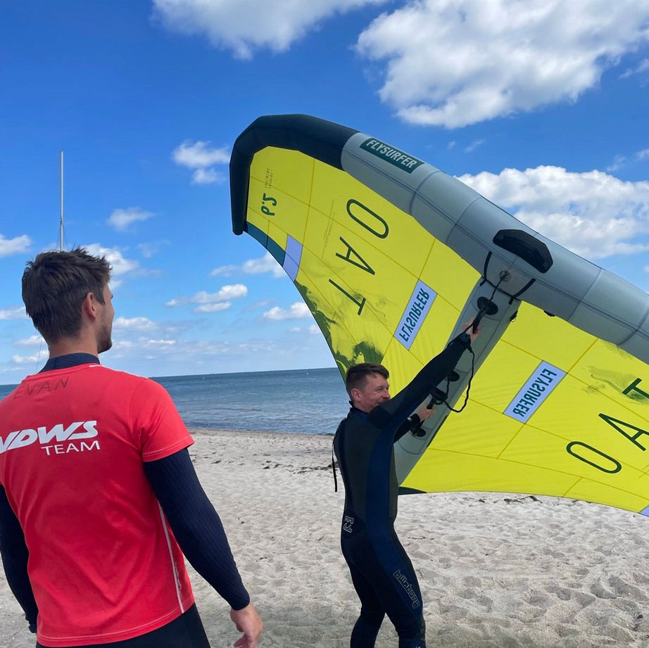 Zwei Männer stehen am Strand, einer der beiden hält das Wing-Segel
