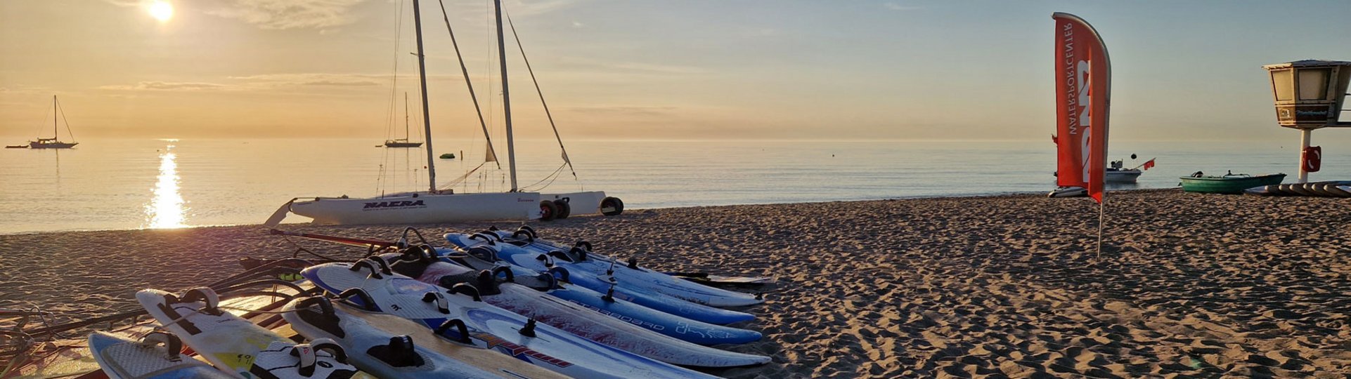 Mehrer Surfbretter liegen zum Trocknen am Strand, im Hintergrund geht die Sonne über dem Wasser unter