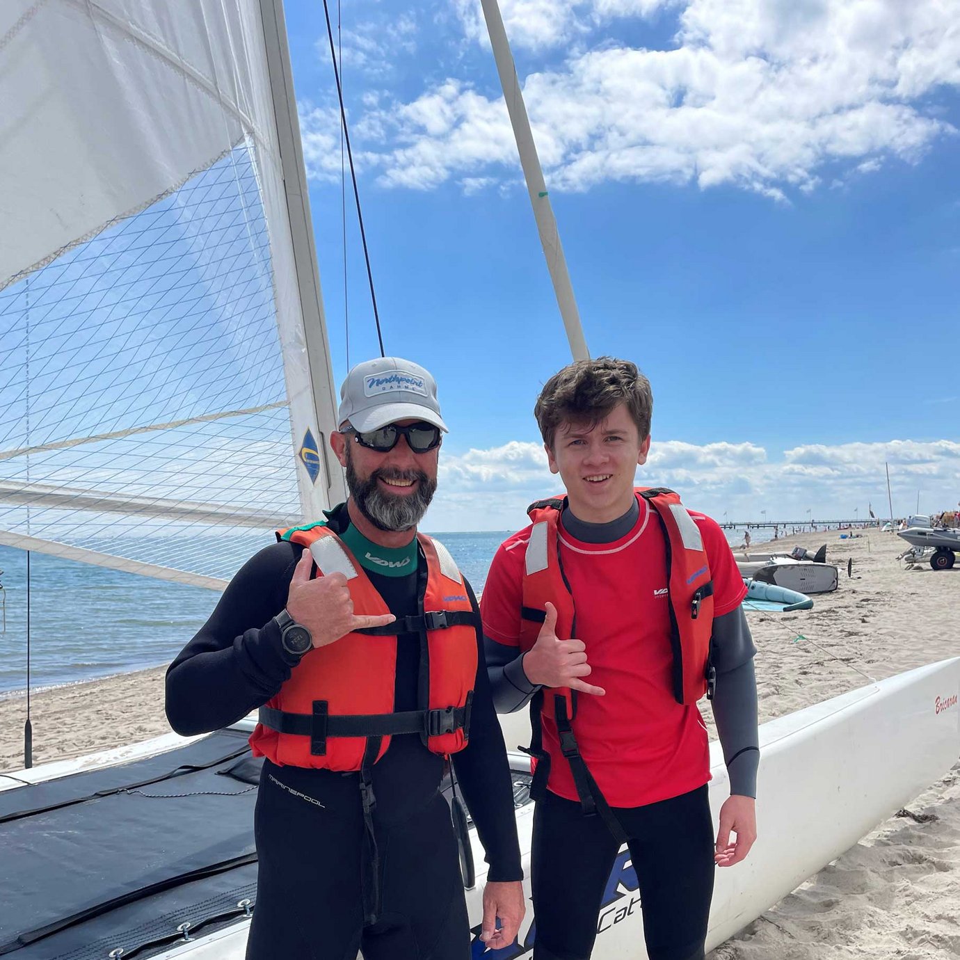Vater und Sohn stehen bei sonnigem Wetter am Strand vor einem Katamaran und lächeln in die Kamera