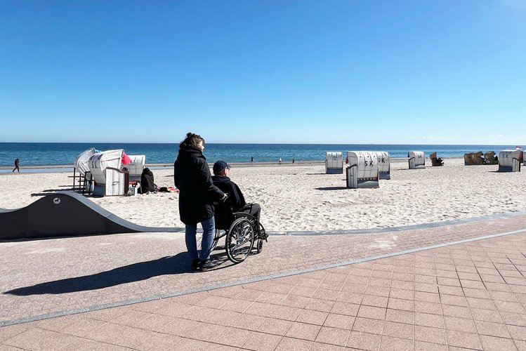 Eine Frau schiebt einen Mann im Rollstuhl bei Sonnenschein über eine Strandpromenade