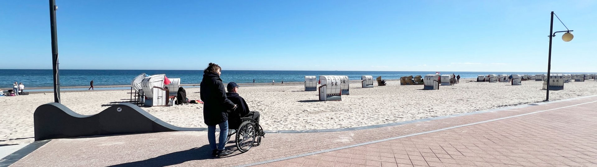 Eine Frau schiebt einen Mann im Rollstuhl bei Sonnenschein über eine Strandpromenade